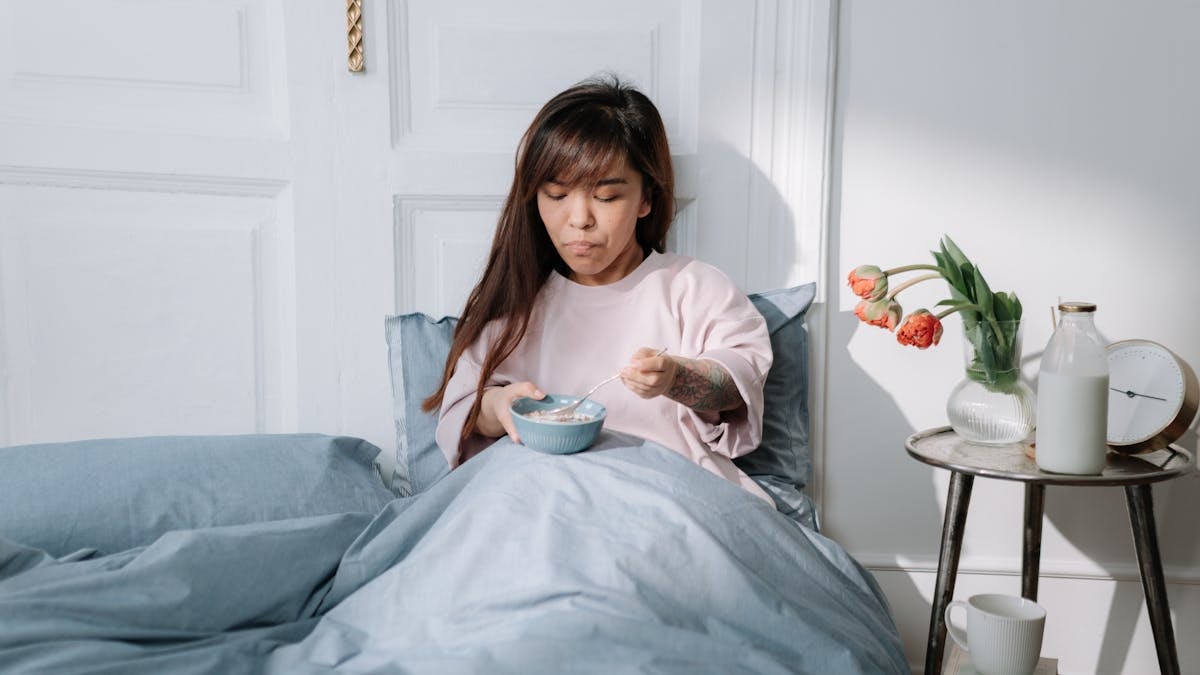 Young woman enjoys breakfast in bed with a bowl of cereal in a cozy bedroom setting.